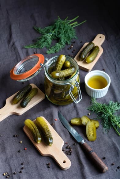 Draufsicht-Flatlay eines kompletten Gewürzgurken-Arrangements mit Einmachgläsern, Cornichons auf einem Holzbrett, Dill, Pfefferkörnern und geschnittenen Gewürzgurken auf dunklem Leinen