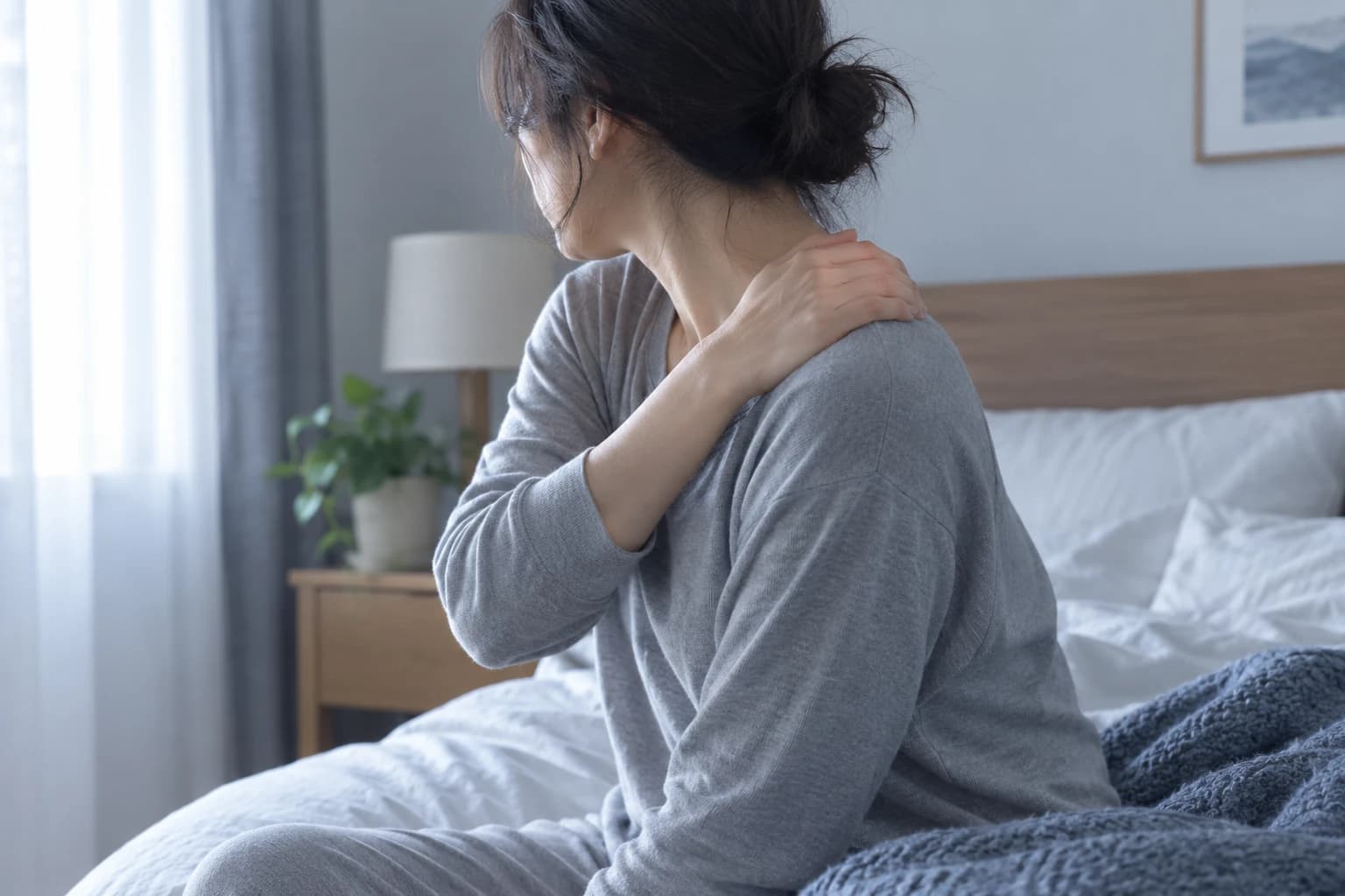 A tired adult sitting on the edge of a bed in early-morning light, rubbing a sore shoulder