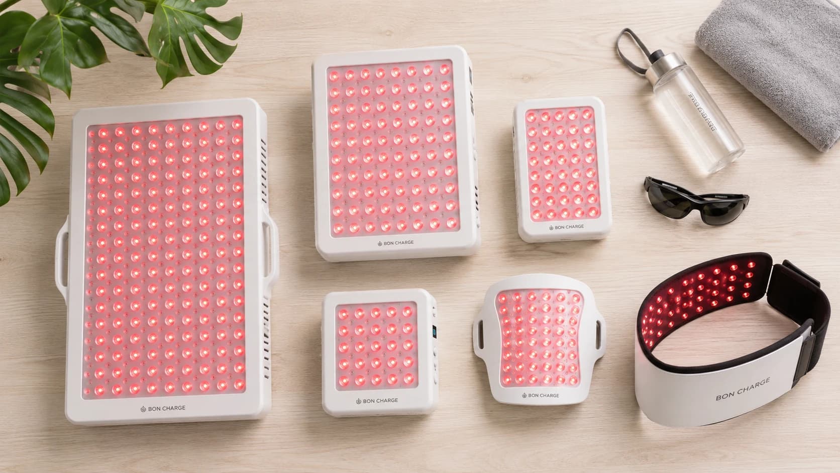 Flatlay of six red light therapy devices for muscle recovery, including panels of various sizes and a wearable wrap on a light wood surface